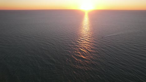 Drone-clip-over-open-sea-at-beautiful-orange-sunset,-moving-towards-a-boat-moving-right-to-left-leaving-ripples-across-the-water