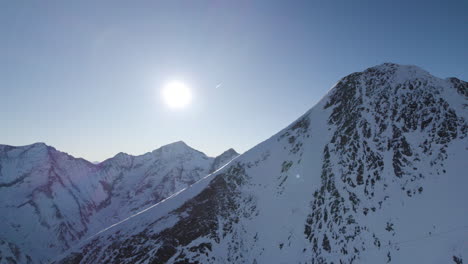 Drone-panning-around-Austrian-mountain-peak-revealing-bright-sun-with-mountain-range-in-background
