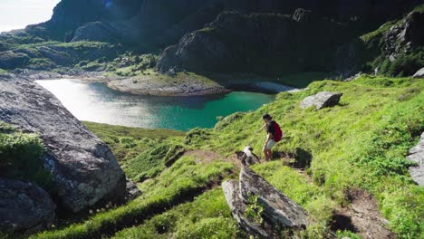 Female-Trekker-Walking-Downhill-With-Pet-Dog-With-Stunning-Scenery-Of-Turquoise-Blue-Mountain-Lake-In-Lovund,-Norway