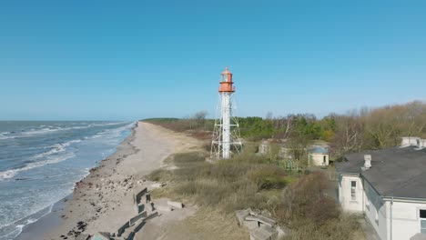 Aerial-establishing-view-of-white-colored-Pape-lighthouse,-Baltic-sea-coastline,-Latvia,-white-sand-beach,-large-waves-crashing,-sunny-day-with-clouds,-wide-ascending-drone-shot-moving-forward