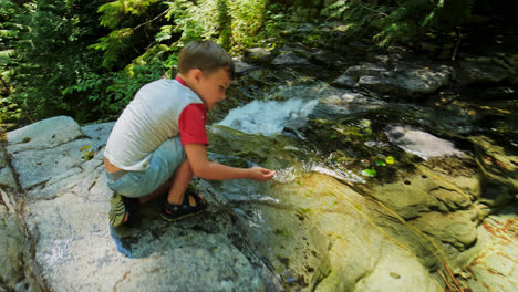 Young-Boy-Kid-Crouched-Down-Next-to-Waterfall-on-Top-of-Rocky-Cliff-in-Nature-Park,-British-Columbia-Canada