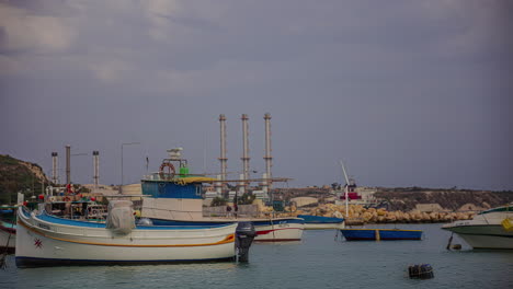 Pequeños-Barcos-Privados-Amarrados-Cerca-De-La-Costa-De-Malta-Con-Edificios-Industriales-En-El-Fondo