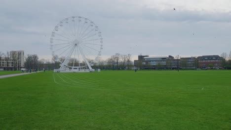 Pan-across-white-Ferris-wheel-behind-green-grass-track-in-Cambridge-England