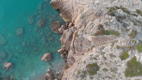 drone-captures-birds-eye-of-rocky-cliff-edge,-as-camera-soars-over-rugged-landscape-of-blue-water-and-white-stone-wall