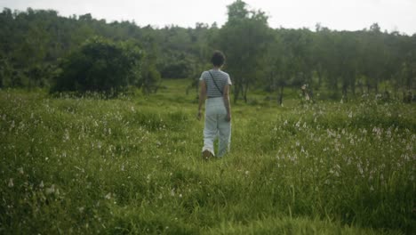 Young-woman-walking-through-deep-green-grass-and-trees,-looking-along-flat-land,-filmed-at-waist-height