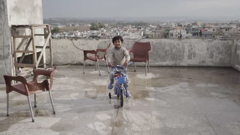Un-Joven-Paquistaní-Asiático-De-Cinco-Años-Está-Montando-En-Bicicleta-Bajo-La-Lluvia-En-Un-Techo