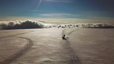 Aerial-view-of-a-person-doing-stunts-on-a-snowmobile,-on-a-glacier-in-Iceland,-over-the-clouds,-on-a-sunny-day