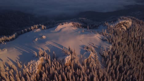 Experience-the-enchantment-of-a-winter-cabin-atop-a-snow-covered-mountain,-a-captivating-hideaway-amidst-a-picturesque-sunrise,-amazing-trees-drone-view