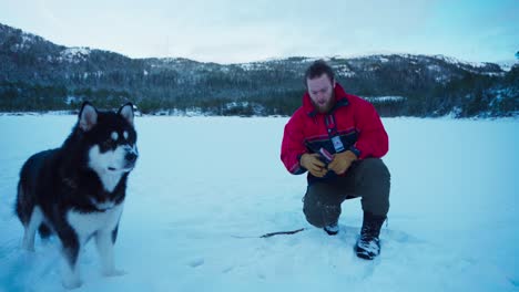Man-And-His-Alaskan-Malamute-Pet-Dog,-Ice-Fishing-In-A-Frozen-Lake