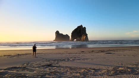 Female-photographer-with-camera-walking-on-sandy-beach-at-famous-picturesque-spot-of-Wharariki-Beach,-New-Zealand