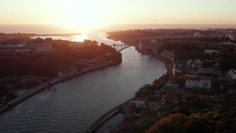 Drone-flyover-view-of-river-with-bridge-and-city-during-sunset-on-horizon,-Porto,-Portugal