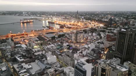 Aerial-flyover-view-Montevideo-downtown-high-rise-cityscape-towards-shipyard-docks-illuminated-at-night
