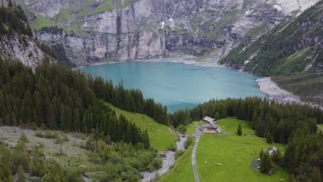 Aerial-drone-view-wooden-cabin-chalet-farm,-Cattle-on-green-alpine-meadow-surrounded-by-alp-mountains,-pine-tree-forest-overlook-turquoise-emerald-glacier-lake-Oeschinensee-in-Kandersteg,-Switzerland