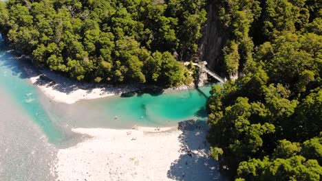 Bird's-Eye-View-Of-The-Beautiful-Blue-Pools-Of-Makarora-River-In-West-Coast,-New-Zealand-With-Tourists-Enjoying-On-The-White-Sand-And-Swing-Bridge-At-Summer---descending-drone-shot