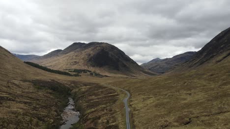 Aerial-view-of-winding-highway-by-a-flowing-river-in-a-valley-surrounded-by-mountains-in-Scotland