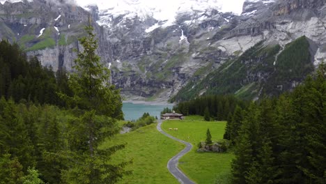 Vista-Aérea-Ascendente-Cabaña-De-Madera-Aislada-Chalet-Granja-Sola-En-Verde-Pradera-Alpina-Rodeada-De-Montañas-Alpinas,-Pinos-Con-Vistas-Al-Lago-Glaciar-Azul-Turquesa-Oeschinensee-En-Kandersteg,-Suiza
