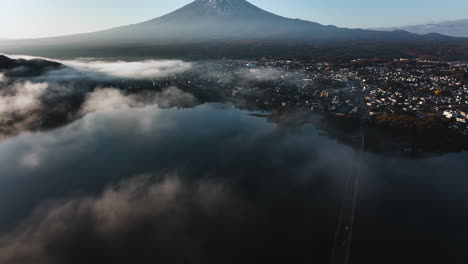 Low-hanging-clouds-above-Lake-Kawaguchi,-revealing-the-Fujikawaguchiko-village-and-Mt-Fuji,-sunrise-in-Japan---Aerial-view
