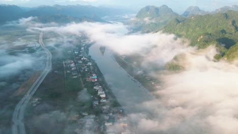 Drone-shot-of-small-town-by-green-river-beneath-dreamy-white-cloud-floating-by-limestone-mountain-at-sunrise