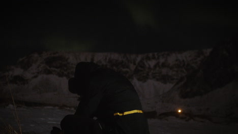 Lone-man-camps-in-snowy-fjord,-meditates-under-night-sky-of-aurora,-Lofoten