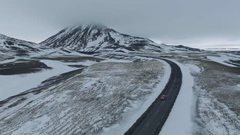 Aerial-View-of-Red-Car-on-Wet-Icelandic-Ring-Road-and-Snow-Capped-Volcanic-Hills