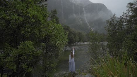 Lady-in-long-dress-standing-on-rock-shore-of-river-with-distant-waterfalls,-magical-scene