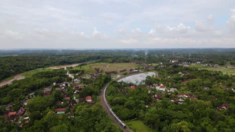 Aerial-birds-eye-shot-of-ride-train-between-green-area-of-Indonesia-near-dirty-brown-river-in-small-village