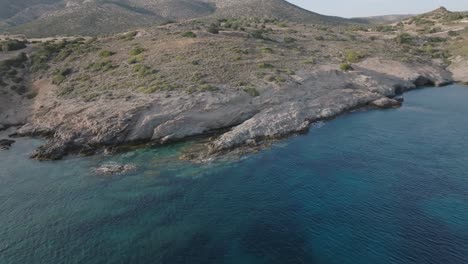 Aerial-backwards-shot-of-clear-shoreline-with-rocks-and-mountain-landscape-of-Greece-in-background