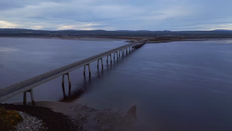 Drone-glides-gracefully-towards-the-Dornoch-Firth-Bridge-during-the-enchanting-sunset-blue-hour,-revealing-a-breathtaking-view-of-the-bridge-spanning-over-the-glistening-ocean