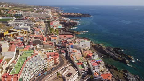 Revealing-wide-view-of-coastal-city-Puerto-de-Santiago,-featuring-beautiful-colourful-hotels-and-residential-buildings-along-coast-in-Santa-Cruz-de-Tenerife,-Canary-islands,-Spain