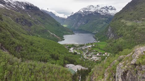 Mighty-Geiranger-Fjord-seen-from-Flydalsjuvet-viewpoint---Forward-moving-aerial-suring-springtime-with-lush-green-forest-and-snow-capped-mountain-peaks