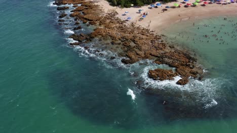 Bird's-eye-aerial-drone-view-of-the-popular-tropical-Coquerinhos-beach-covered-in-umbrellas-with-tourists-swimming-in-a-natural-pool-from-a-reef-blocking-small-waves-in-Conde,-Paraiba,-Brazil