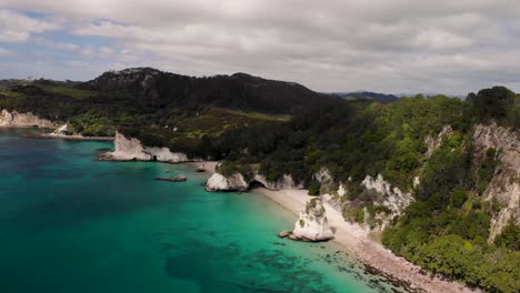 Cathedral-Cove-at-Coromandel-Peninsula-aerial-orbit-reveal-of-cave,-white-sandy-beach,-cliffs-and-forest-landscape,-New-Zealand-seaside