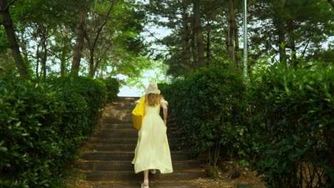 Woman-in-yellow-dress-walking-up-stairs-through-trees-carrying-bag,-from-behind