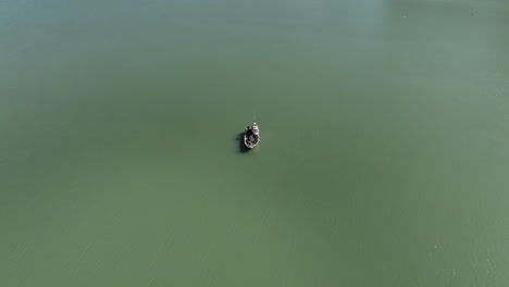 Aerial-view-circling-tiny-fishing-boat-floating-on-shimmering-sunlit-emerald-green-water