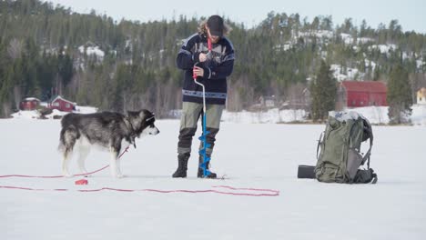 Hund-Beobachtet-Den-Kerl,-Der-Ein-Loch-Auf-Dem-Zugefrorenen-See-Bohrt---Eisfischen