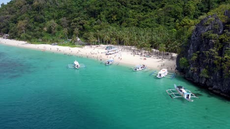 Aerial:-Filipino-Outrigger-Tour-A-Boats-unloading-Island-hopping-tourists-on-Tropical-Seven-Commandos-Beach,-turquoise-clear-water-on-white-sand,-Palm-trees-and-lush-green-limestone-mountains