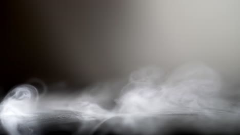 Slow-motion-fog-smoke-wispy-spins-from-side-spread-over-leather-surface,-grey-background