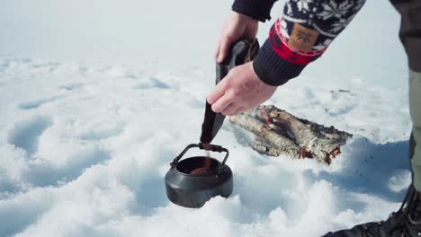 Man-Pouring-Coffee-Grounds-On-Kettle-Near-The-Burning-Wood-In-The-Snow