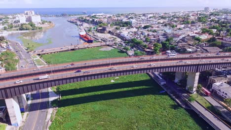 Autoverkehr-über-Der-Brücke-Puente-Ramon-Matias-Mella-über-Den-Rio-Ozama,-Verstopft-Mit-Invasiver-Wasserhyazinthe,-Luftaufnahme