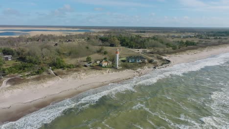 Aerial-establishing-view-of-white-colored-Pape-lighthouse,-Baltic-sea-coastline,-Latvia,-white-sand-beach,-large-waves-crashing,-sunny-day-with-clouds,-wide-drone-orbit-shot