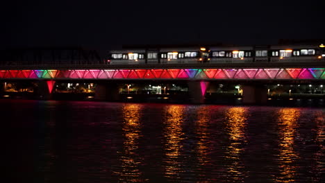 Tranvía-Sobre-El-Puente-De-Agua-Por-La-Noche-Con-Luces-De-Colores