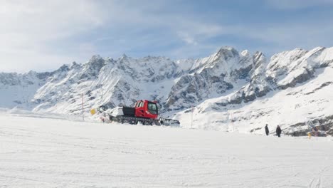 Wide-shot-of-snow-plow-drive-on-a-slope-in-Cervinia-ski-resort-with-mountain-range-in-background