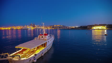 Nächtlicher-Zeitraffer-Von-Valletta-Malta-Im-Hintergrund-Mit-Einem-Boot-Und-Der-Bucht-Im-Vordergrund