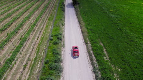 Antena-Siguiendo-Un-Coche-Rojo-En-Una-Carretera-Rural-Por-Tierras-De-Cultivo-Verdes-En-Toscana
