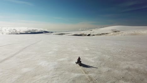 Aerial-view-of-a-person-riding-a-snowmobile-on-a-white-glacier-in-Iceland,-over-the-clouds,-on-a-sunny-day