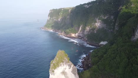 Aerial,-flying-above-hidden-cove-sandy-beach-and-diamond-shaped-rock-formation-along-lush-steep-limestone-coastline-cliffs-of-Nusa-Penida-Island-on-a-misty-morning