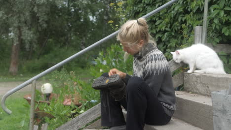 Pretty-Blonde-Nordic-Woman-Greasing-Her-Leather-Hiking-Boots-on-a-Farm,-Girl-Cleaning-her-Shoes-on-a-Staircase,-Finland