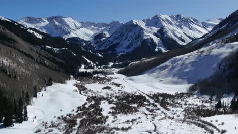 Above-on-a-bluebird-sunny-day-spring-winter-Colorado-beautiful-mountain-views-and-peaceful-stream-Ashcroft-Maroon-Bells-Aspen-Colorado-iconic-scenery-aerial-forward-pan-reveal-motion