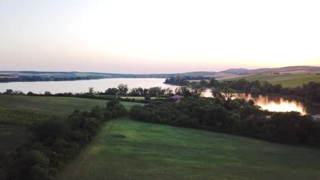 Aerial-View-Green-Meadow-Near-The-Calm-Lake-At-Dusk