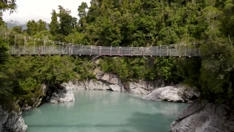 Beautiful-aerial-reveal-of-swing-bridge-above-glacial-pristine-water-of-Hokitika-Gorge-surrounded-by-lush-green-bush